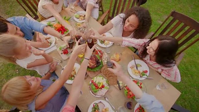 家人和朋友聚集在餐桌旁，举起玻璃杯和瓶子敬酒并碰杯。大家庭花园派对庆典。升降自上而下的相机拍摄。