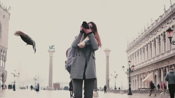 快乐的女摄影师用相机拍摄威尼斯的鸽子群在老城广场微笑慢动作的照片。