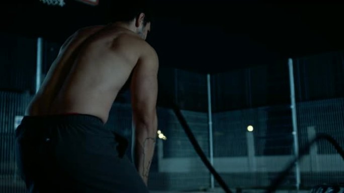 一个强壮的肌肉健康的光着膀子的年轻人用战斗绳做练习的背景。他正在一个有围栏的室外篮球场里锻炼身体。居