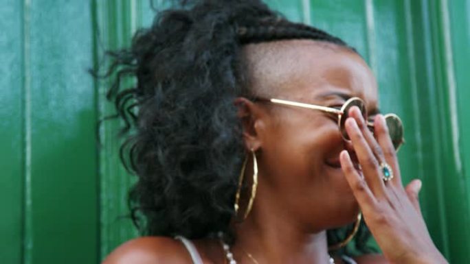 戴着墨镜的年轻黑人妇女在笑，头和肩膀上靠在绿色的墙上