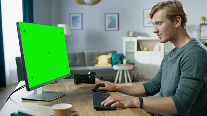 英俊的年轻摄影师坐在舒适公寓的办公桌前，在绿色模拟屏幕个人电脑上工作。