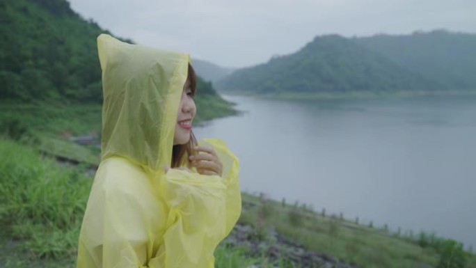 年轻的亚洲女子穿着雨衣站在湖边，在雨中玩耍时感到快乐。生活方式女性在雨天享受和放松。
