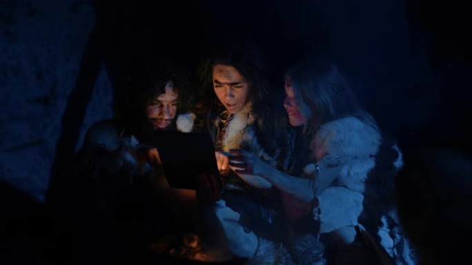 穿着兽皮的史前原始狩猎采集者部落在夜间在洞穴中使用数字平板电脑。尼安德特人或智人家庭浏览互联网、观看