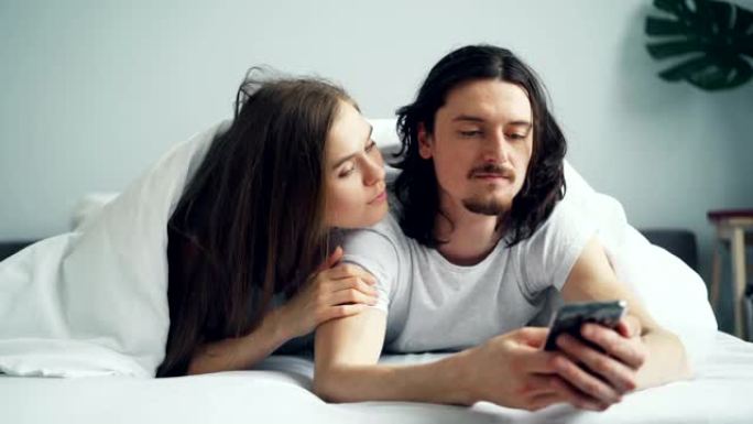 男人在手机上聊天，而漂亮的妻子在床上咬着耳朵笑