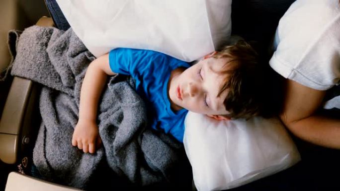 4-6岁的高加索小男孩在长途飞机飞行中睡在舒适的白色枕头上的俯视图照片。