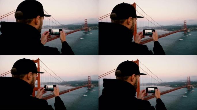 背面看黑色连帽衫和帽子的年轻大胡子男人拍摄旧金山日落金门大桥的智能手机照片。