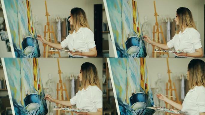 金发女孩画家在工作室里工作，用画笔和蛋彩画画在画布上描绘海洋景观和船只，看着画面并享受她的职业。