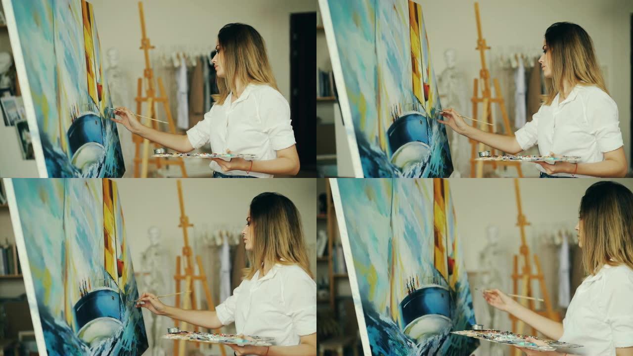金发女孩画家在工作室里工作，用画笔和蛋彩画画在画布上描绘海洋景观和船只，看着画面并享受她的职业。