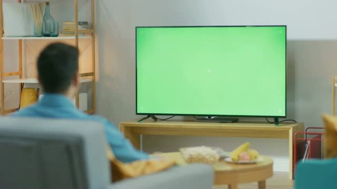 快乐的家伙坐在客厅的椅子上，看着绿色色度键屏幕，下班后放松。男人在舒适的房间里看体育比赛，新闻，电视