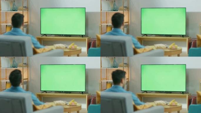 快乐的家伙坐在客厅的椅子上，看着绿色色度键屏幕，下班后放松。男人在舒适的房间里看体育比赛，新闻，电视