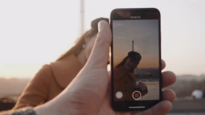 男性的手正在制作智能手机视频，在度假慢动作时，快乐微笑的女人在日落埃菲尔铁塔附近摆姿势。