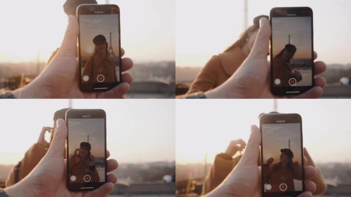 男性的手正在制作智能手机视频，在度假慢动作时，快乐微笑的女人在日落埃菲尔铁塔附近摆姿势。