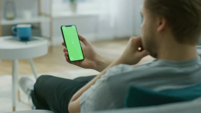 家里的年轻人使用绿色模拟屏幕智能手机。她坐在他舒适客厅的沙发上。肩膀上的相机拍摄