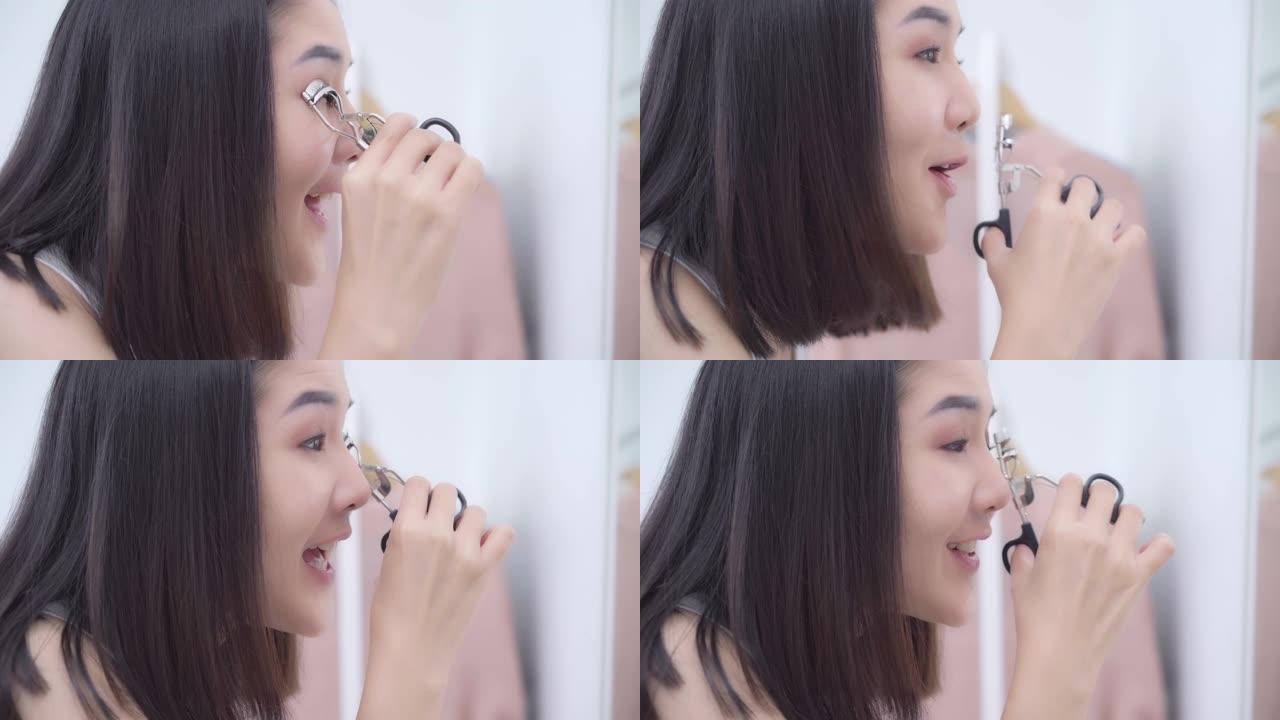 美丽的亚洲女人在前镜中使用睫毛夹化妆，快乐的女性使用美容化妆品来改善自己，准备在家卧室工作。生活方式