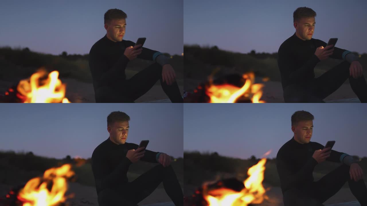 当太阳落在她身后时，男子坐在海滩上的篝火旁，用手机坐在冲浪板上。