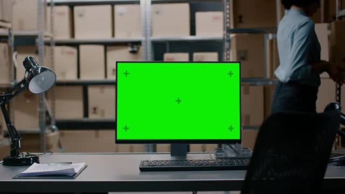 在带有绿色模拟屏幕显示的仓库计算机中，站在桌面上。在背景成排的货架上，装满了准备好发货的纸板箱和包裹