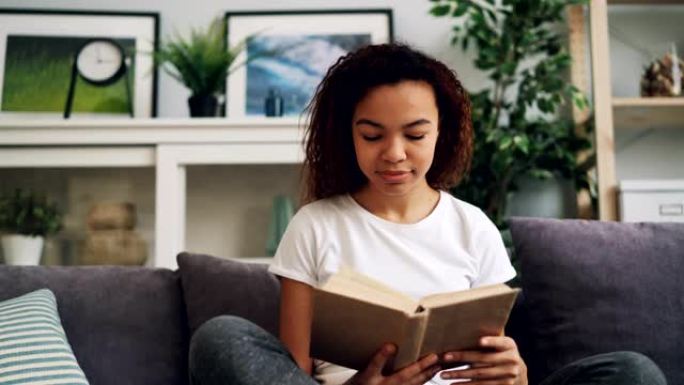 迷人的年轻非洲裔美国妇女独自坐在沙发上看书，享受现代文学。爱好、青年和室内概念。