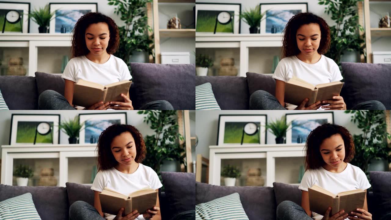 迷人的年轻非洲裔美国妇女独自坐在沙发上看书，享受现代文学。爱好、青年和室内概念。