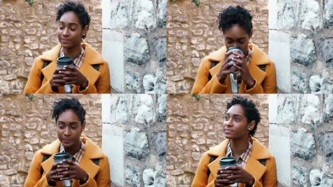 千禧一代黑人妇女穿着黄色外套靠在小巷里的石墙上喝外卖咖啡，特写