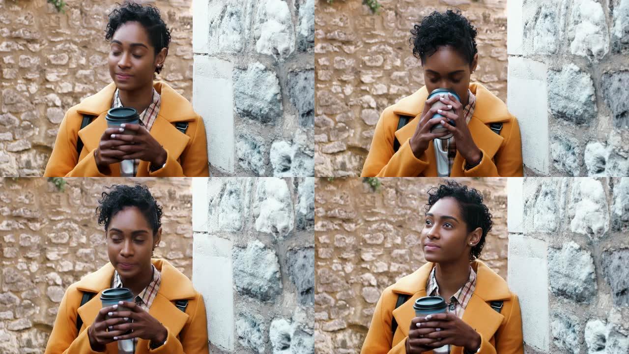 千禧一代黑人妇女穿着黄色外套靠在小巷里的石墙上喝外卖咖啡，特写