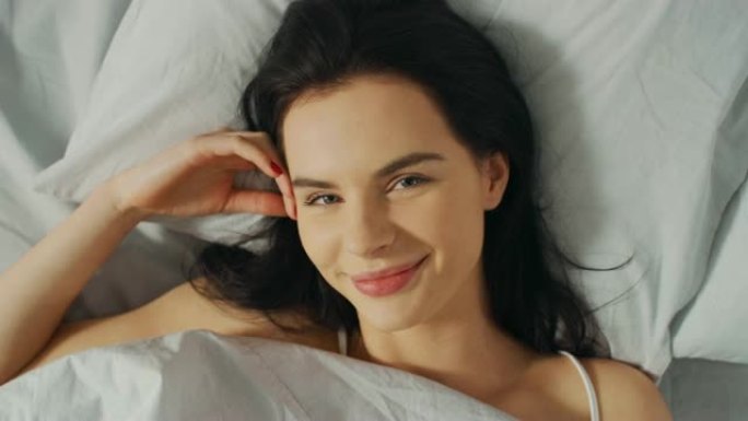 美丽的年轻黑发女人的肖像迷人地在她的床上醒来，性感地微笑着，她的脸被温暖的早晨的阳光照亮。甜美温暖。