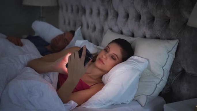 女人躺在床上检查手机，而男人睡在她旁边