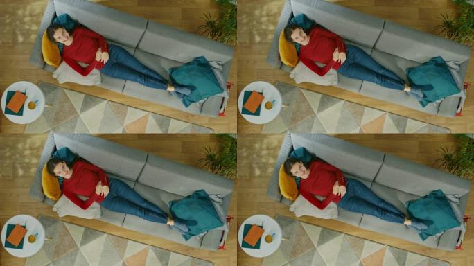 穿着红色外套和蓝色牛仔裤的年轻女孩躺在带枕头的沙发上。看着上面微笑。舒适的客厅，配有现代室内地毯、植