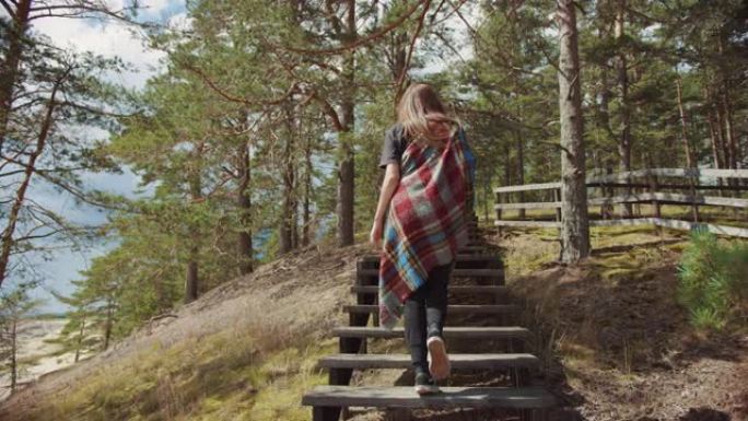 年轻漂亮的女孩走在松林边缘的木制楼梯上，从美丽的蓝色海景出发。温暖阳光明媚的日子与大自然。她裹着格子