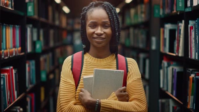大学图书馆学习: 一个聪明美丽的黑人女孩的肖像，拿着学习教科书微笑着看着相机。真实的学生为课堂作业、