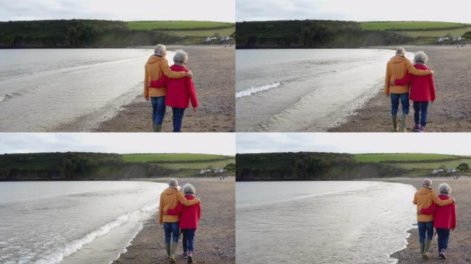 在冬季海滩度假中，爱的高级夫妇手牵着手沿着海岸线行走的无人机镜头从慢动作的背后观看