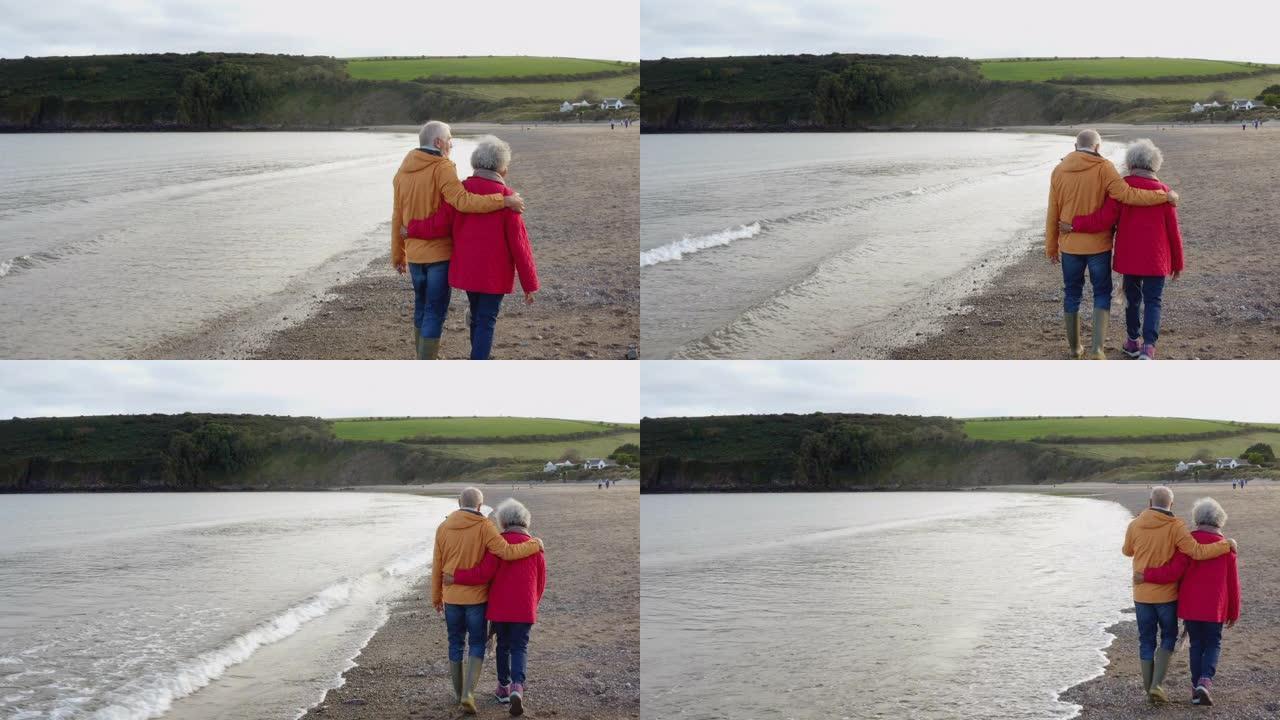 在冬季海滩度假中，爱的高级夫妇手牵着手沿着海岸线行走的无人机镜头从慢动作的背后观看