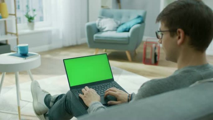 肩膀上的镜头: 家里的年轻人在带有绿色模拟屏幕的笔记本电脑上工作。他坐在舒适客厅的沙发上。