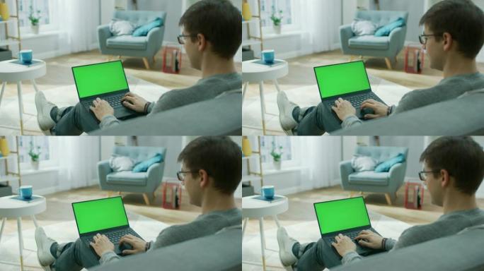 肩膀上的镜头: 家里的年轻人在带有绿色模拟屏幕的笔记本电脑上工作。他坐在舒适客厅的沙发上。