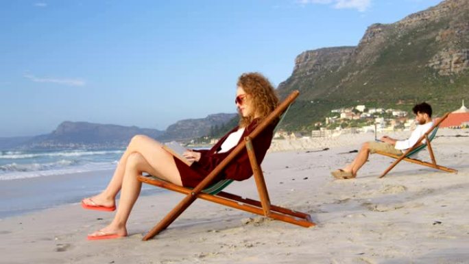 年轻的白人妇女的侧视图，戴着太阳镜坐在躺椅上，在4k海滩看书
