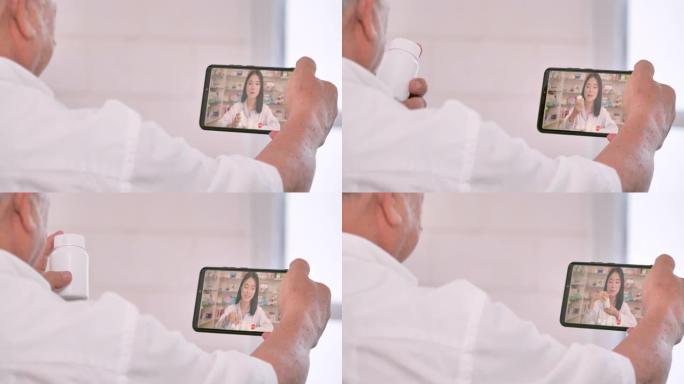 亚洲老年祖父看着药瓶，听亚洲女药剂师在家通过智能手机向视频会议解释咨询医学中的医学。高级健康技术，医
