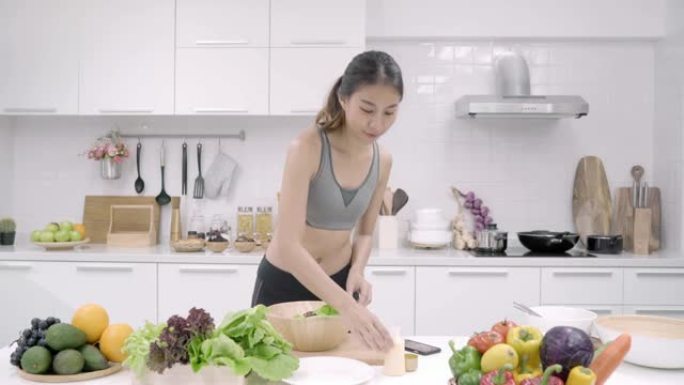 年轻的亚洲妇女在厨房里用手机寻找食谱的同时制作沙拉健康食品，美丽的女性在家中使用蔬菜准备沙拉以保持身