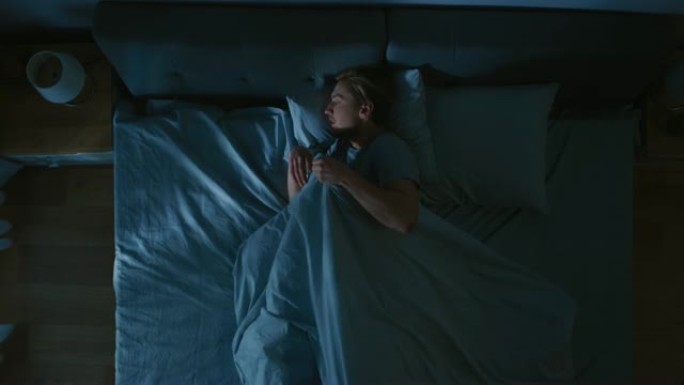 英俊的年轻人晚上舒适地睡在卧室的床上的俯视图。蓝色夜间颜色，寒冷微弱的灯柱光线透过窗户照射。