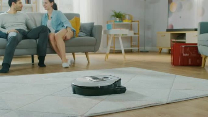 智能机器人真空吸尘器从地毯上吸尘的特写镜头。美丽的夫妇坐在沙发上，在后台聊天。科技家电设备超越了它们