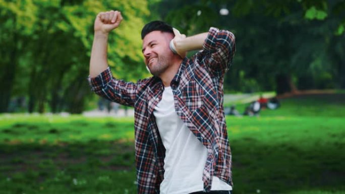 年轻人的慢动作很高兴在阳光明媚的日子里戴着白色耳机听音乐并在绿色城市公园跳舞