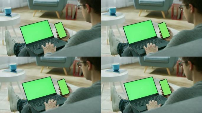 家里的年轻人在带有绿色模拟屏幕的笔记本电脑上工作，同时拿着带有色度键显示的智能手机。他坐在舒适客厅的