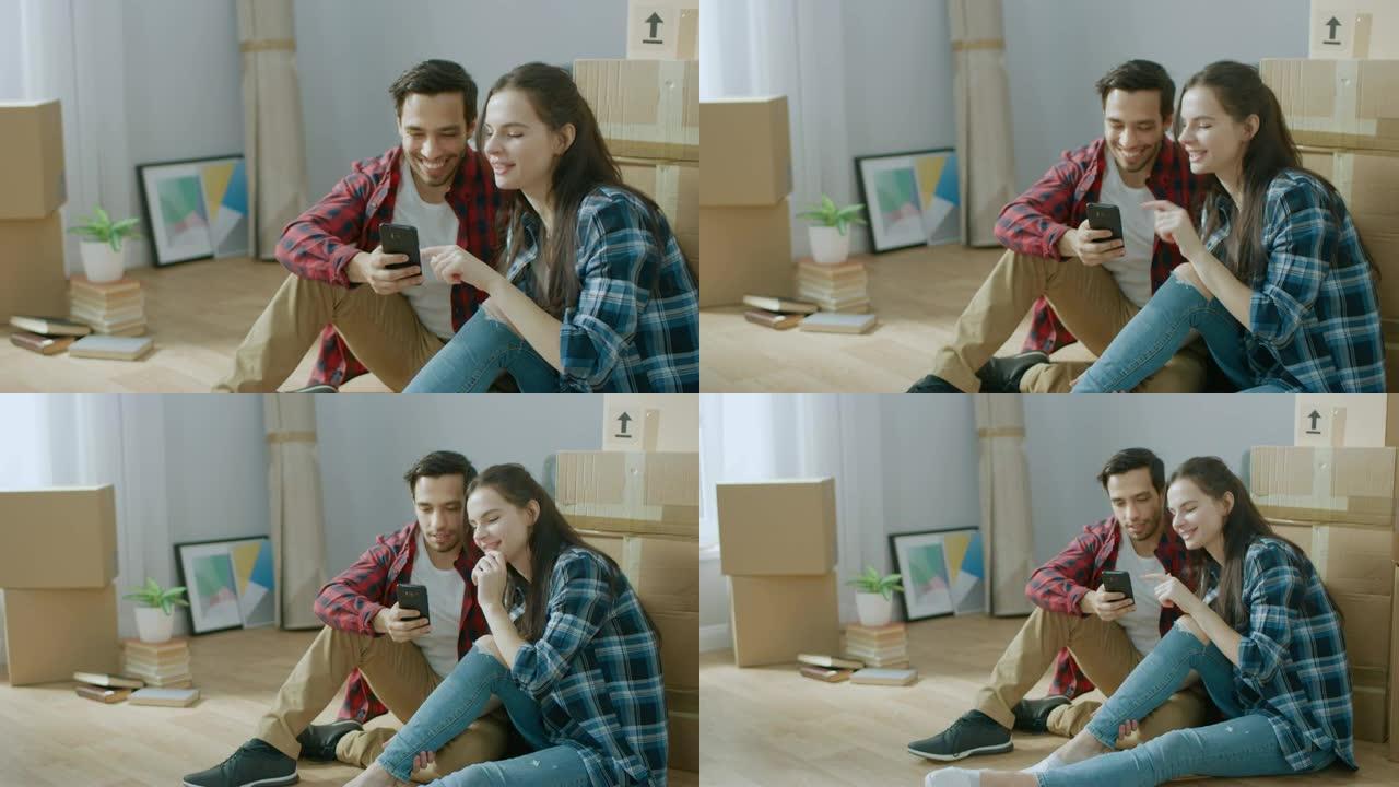 幸福的年轻夫妇坐在新租/购买的公寓的地板上使用智能手机。在现代明亮的甜蜜家庭中打开包装的纸板箱和有盖