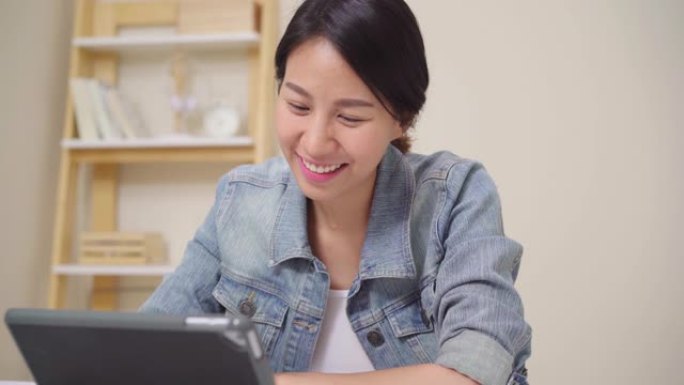 年轻的亚洲女性使用平板电脑检查社交媒体，同时在家客厅的桌子上放松。享受在家的时间概念。