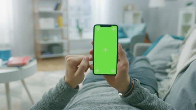 在家中的男子躺在沙发上，使用带有绿色模拟屏幕的智能手机，进行滑动，滚动手势。家伙使用手机，互联网社交