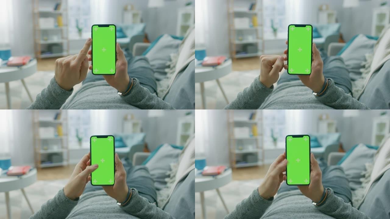 在家中的男子躺在沙发上，使用带有绿色模拟屏幕的智能手机，进行滑动，滚动手势。家伙使用手机，互联网社交