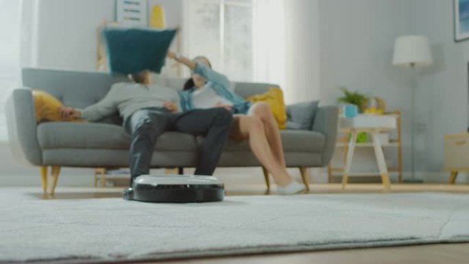 智能机器人真空吸尘器从地毯上吸走污垢的特写镜头。美丽的夫妇坐在沙发上，背景是枕头，观看科技家电设备的