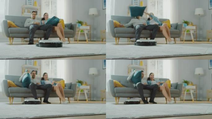 智能机器人真空吸尘器从地毯上吸走污垢的特写镜头。美丽的夫妇坐在沙发上，背景是枕头，观看科技家电设备的