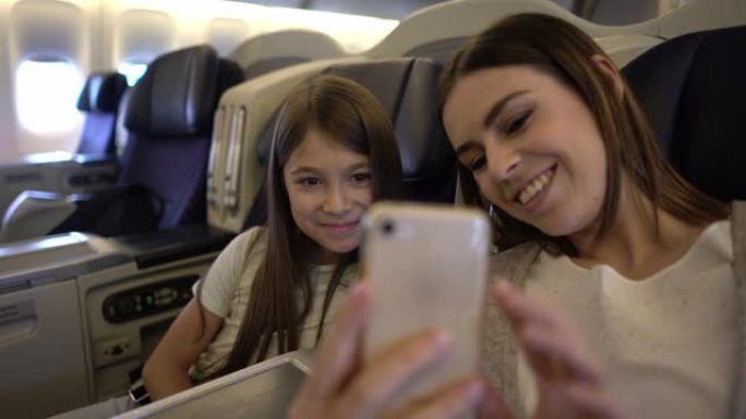 美丽的年轻母亲和女儿一起乘飞机旅行，看着智能手机微笑