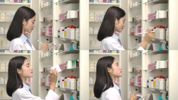 年轻的亚洲药剂师妇女拿着电脑平板电脑，用于检查药房药店盒子上的药物细节。