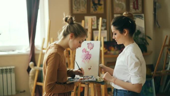 漂亮女人绘画老师正在漂亮的现代工作室教年轻女孩在调色板上混合颜料，在美术课上创造美丽的色彩。人和教育