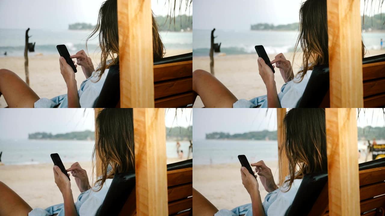 使用智能手机购物应用程序在休闲沙滩椅上放松的年轻女子的后视图照片，拥有令人惊叹的海滨风景。